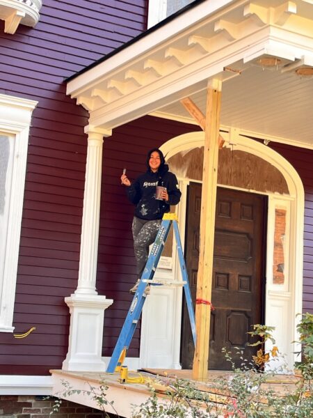 Woman on ladder pauses while painting house to smile at camera.