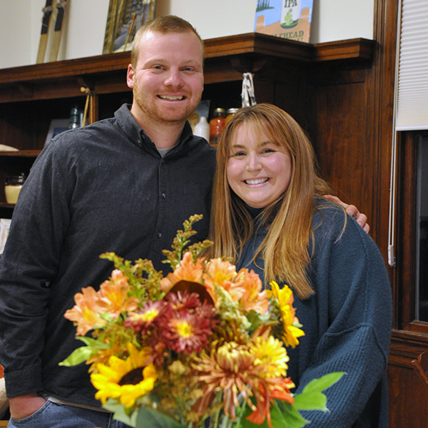Young man and woman stand together behind a vase of colorful fall flowers.
