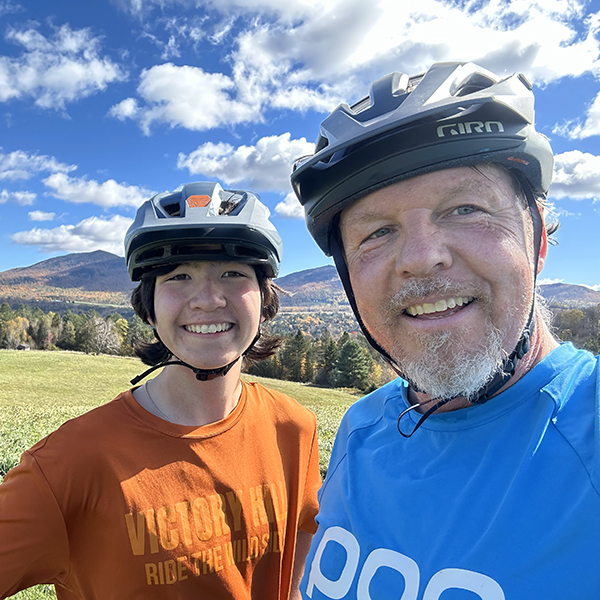 A man and a youth smile while out riding mountain bikes, with Vermont landscape in background.