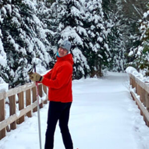 person in red jacket cross country sking across a wooden brige in the snowy woods.