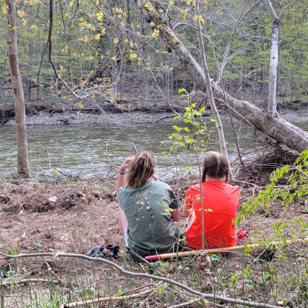 Two girls sit with backs to the camera and watch a small river flow by.