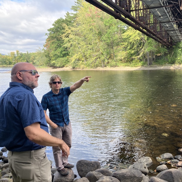 In a summer scene, one man points down river while another man looks on, both standing under a pedestrian bridge that crosses the river.