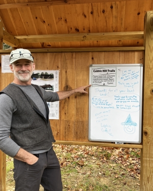 A white man in a ball cap points to messages at a trail kiosk in the fall.