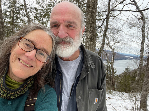 A man and woman standing outdoors with trees and a river in the background smile at the camera. 
