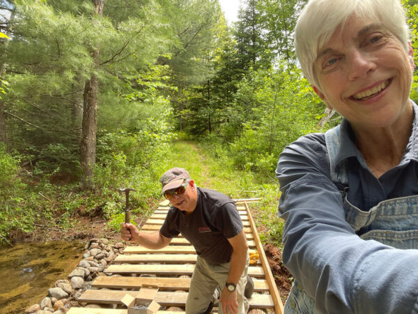 A woman takes a selfie of herself with a man behind her holding a hammer. He is building a small bridge over a creek.