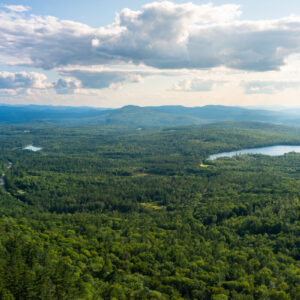 Panoramic view of mountains, forests, and a small lake with partly cloudy skies.