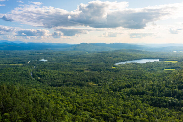 Panoramic view of mountains, forests, and a small lake with partly cloudy skies.